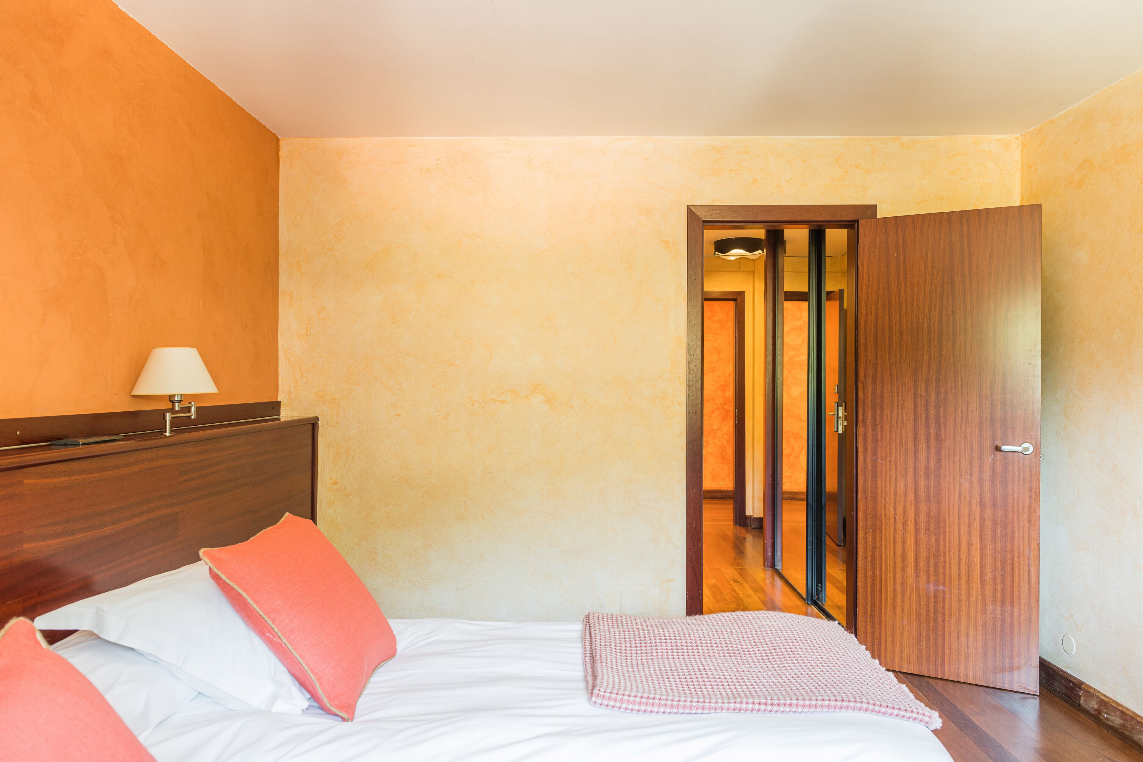 Habitación de hotel con cabecera de madera, cama con ropa de cama blanca y rosa, lámpara de noche y puerta de madera.