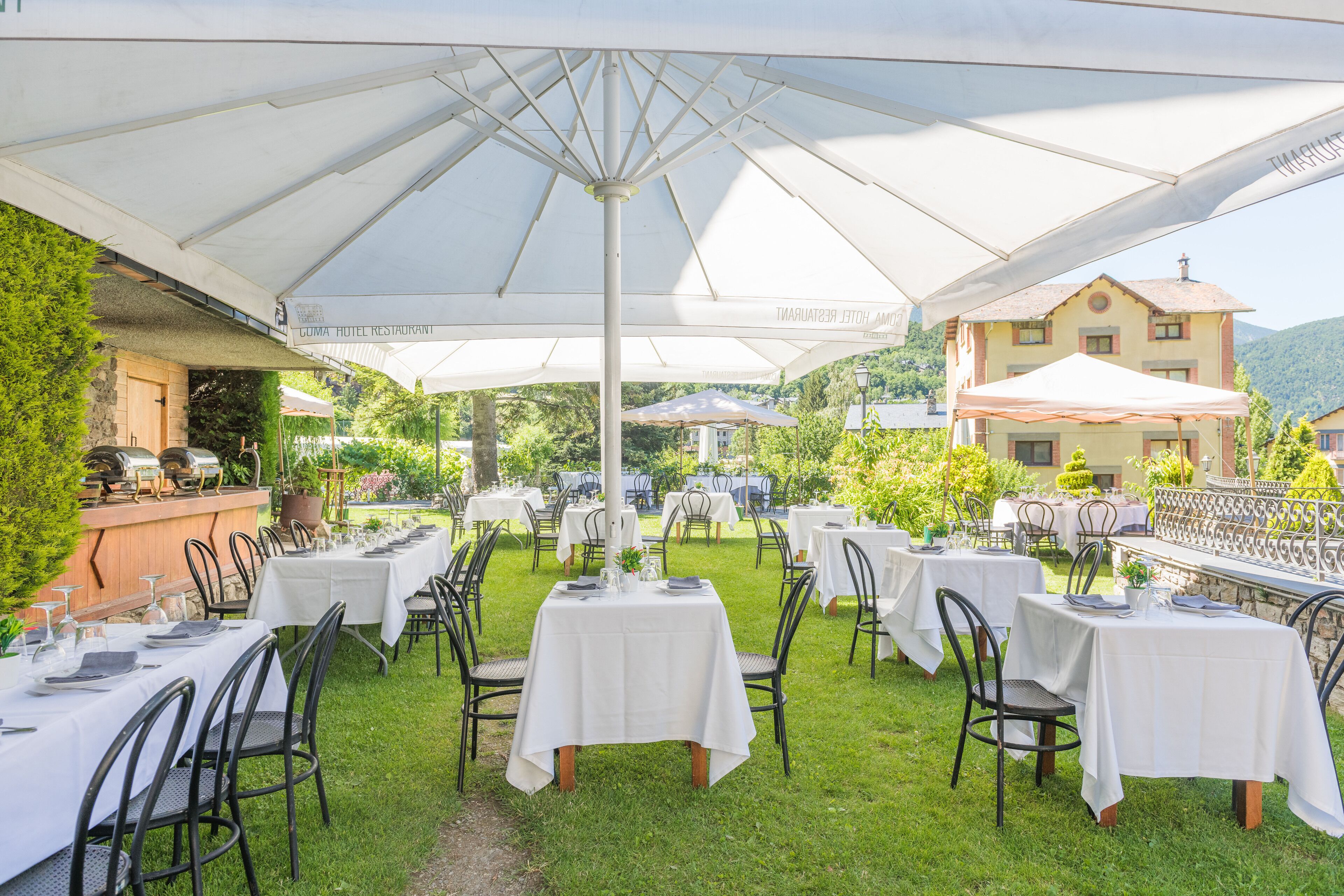 An outdoor dining area with multiple tables and chairs, covered by large umbrellas, set against a backdrop of greenery and buildings.