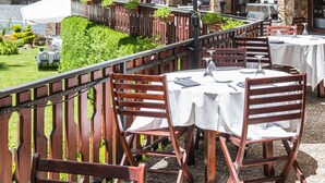 Un área de comedor al aire libre con mesas y sombrillas, barandillas de madera y montañas al fondo.