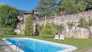 Una piscina con paredes de piedra y césped, rodeada de árboles y una montaña al fondo.
