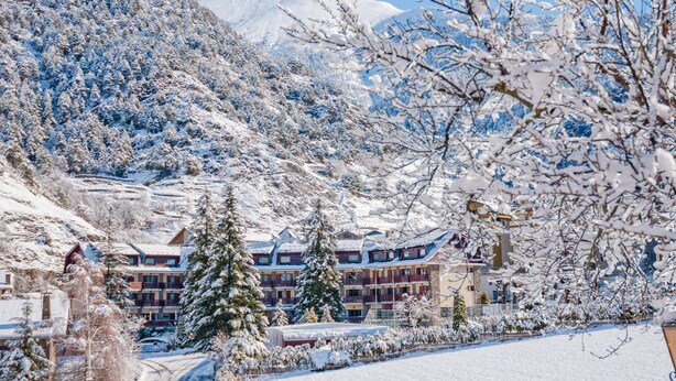 Un complejo turístico en la montaña cubierto de nieve, con un edificio de varios pisos que tiene balcones y una pista de esquí al fondo.