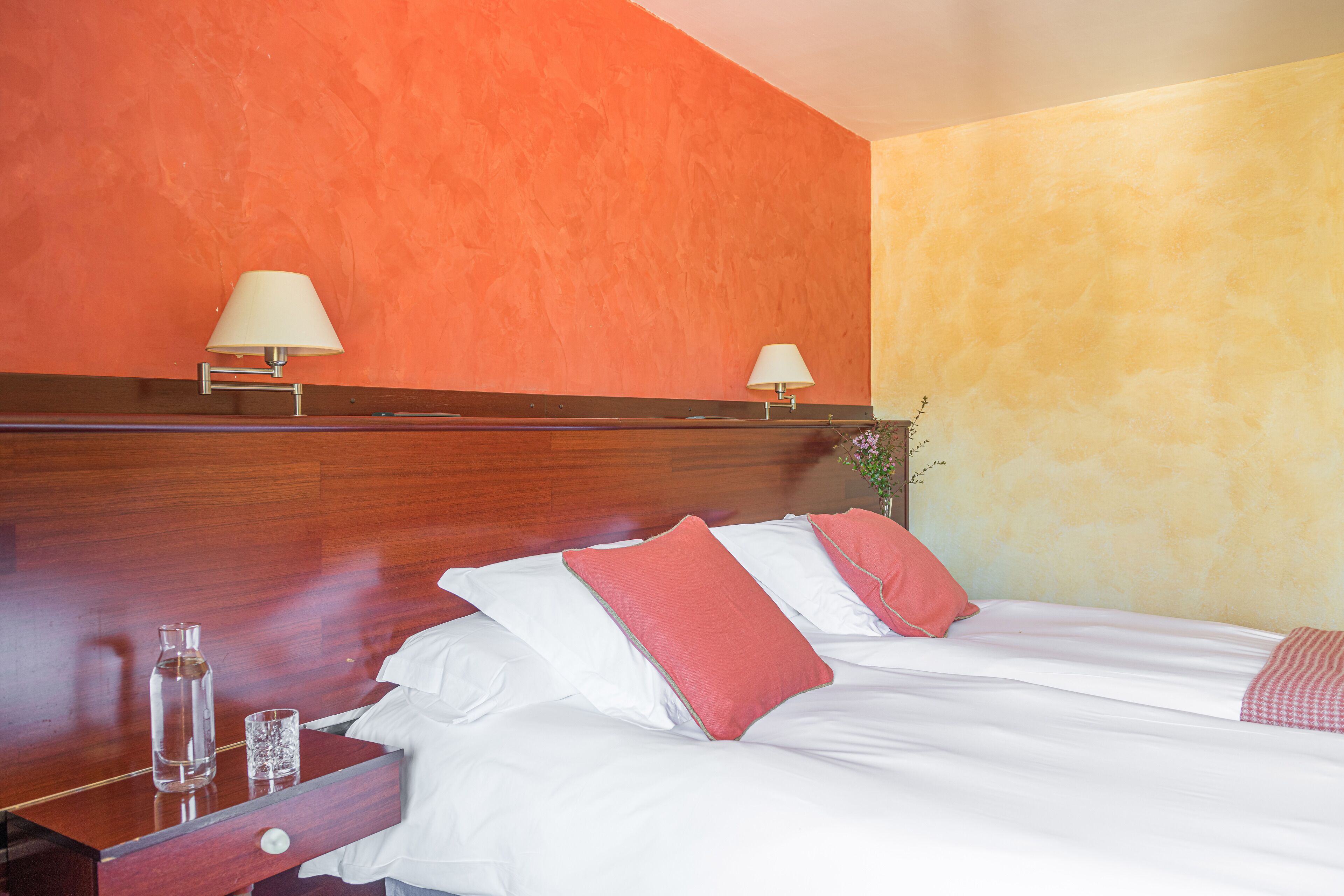 A hotel room with a wooden headboard, two beds with white and red bedding, two bedside lamps, a nightstand with a glass and a bottle, and a wooden dresser.