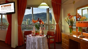 A hotel room with a bed, a table with flowers, a TV, and a view of a cityscape. - Cesars Plaza Hotel (Cochabamba)