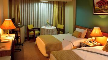 A hotel room with two beds, a desk, a chair, a small table with a vase, and a painting on the wall.