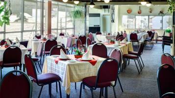 A spacious dining area with tables and chairs, large windows, and hanging plants.