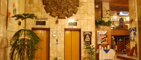 A lobby with a stone wall, two elevators, potted plants, a reception desk, and a religious statue.