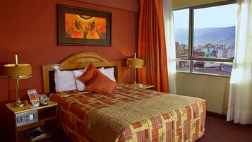 A hotel room with a large bed, two bedside tables with lamps, a framed artwork on the wall, and a window with a city view.