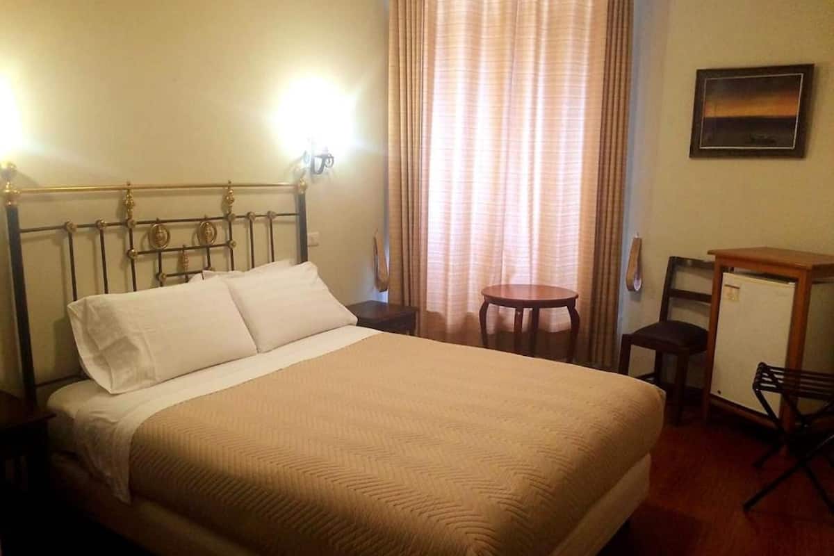 A hotel room with a bed, a desk, a chair, a small table, and a window with curtains.