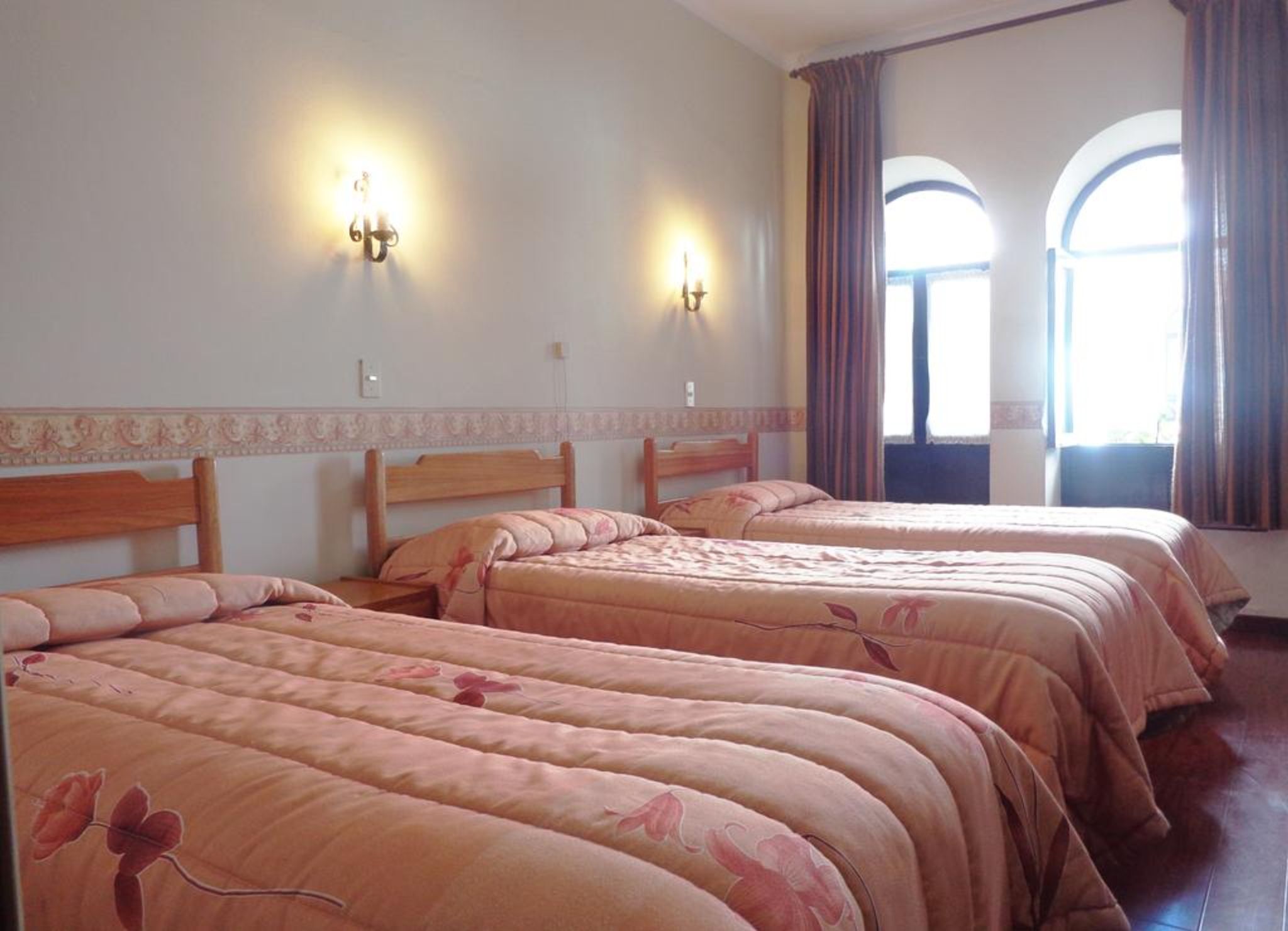 Une chambre d’hôtel avec trois lits, deux appliques murales et de grandes fenêtres en arc surmontées de rideaux.