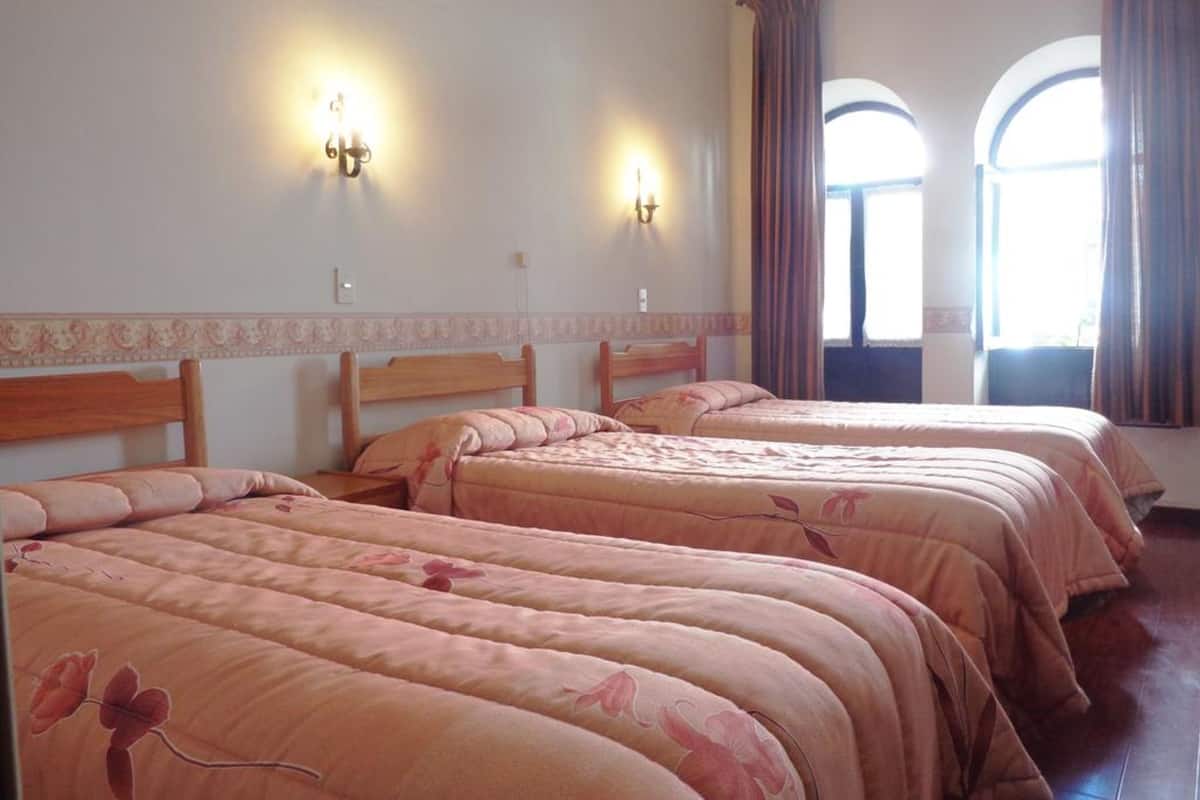 A hotel room with three beds, two wall sconces, and large arched windows with curtains.