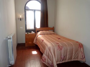 Un dormitorio con una cama individual, una mesita de noche, una lámpara de pared, un radiador y una ventana con cortinas.