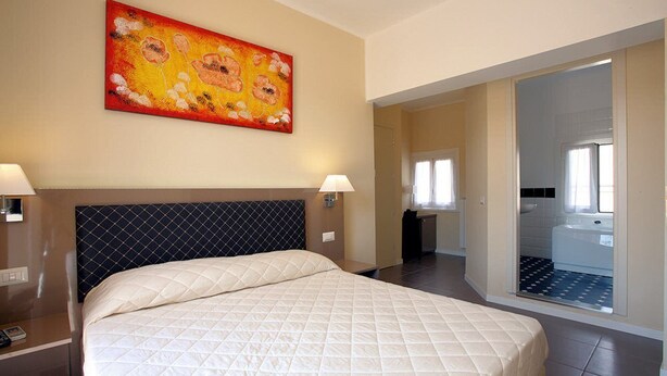 A hotel room with a large bed, a headboard, two bedside lamps, a painting on the wall, and a bathroom visible through an open door. - Hotel Ferton (Milan)