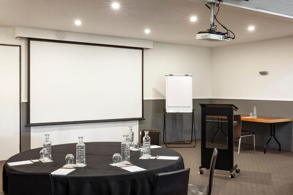 Un salón de conferencias con una pantalla grande, un proyector, una pizarra, una mesa con botellas de agua y un carrito.