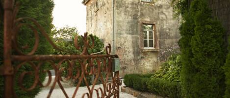 A rustic building with a weathered exterior, a gravel pathway leading to a metal gate, and dense greenery surrounding the property.