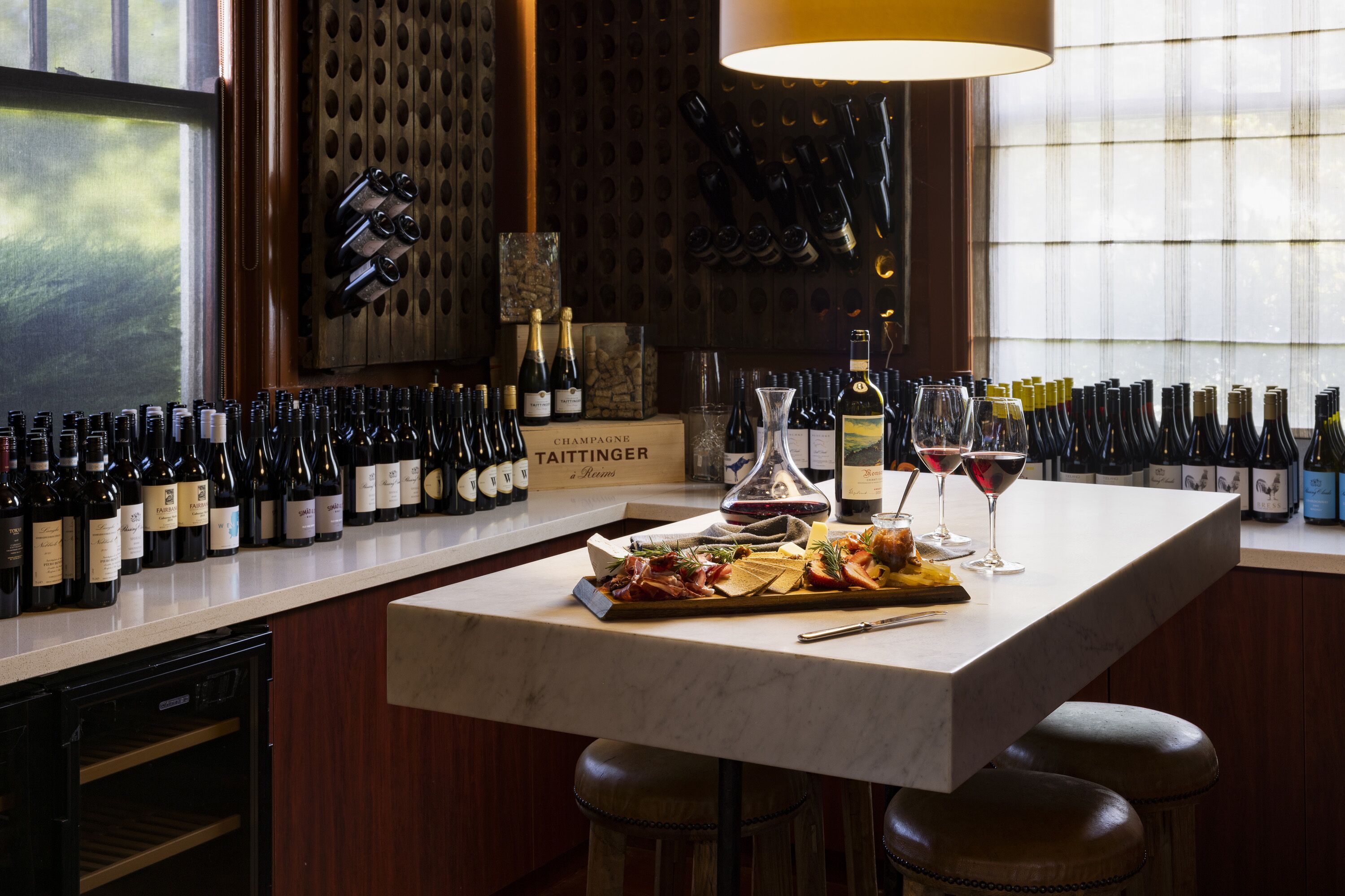 Una zona de bar con mostrador de mármol, botellas de vino, copas y una tabla de embutidos.
