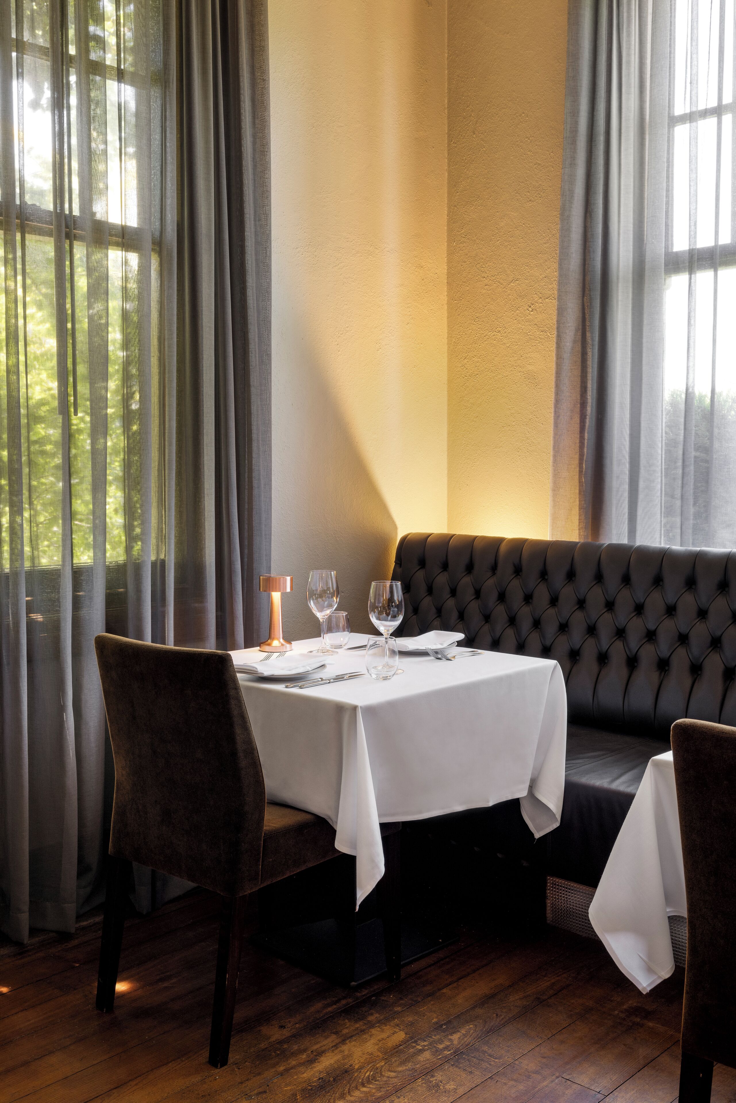 Un coin repas avec une table dressée pour deux personnes, un banc rembourré et une vue dégagée grâce à de grandes fenêtres.