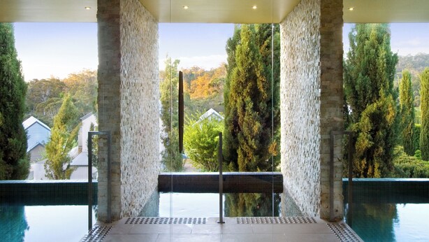A modern indoor pool with stone walls and a clear view of surrounding trees and houses. - Mineral Springs Hotel (Hepburn Springs)