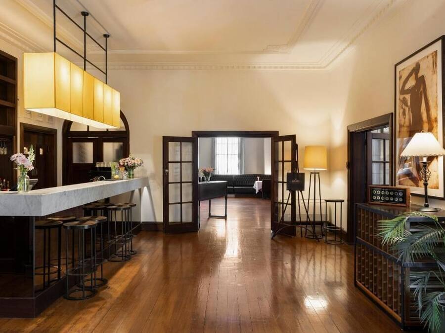 Zona de bar con una barra larga, taburetes y una lámpara colgante grande. Se aprecia una zona de estar con un sofá y una mesa al fondo.