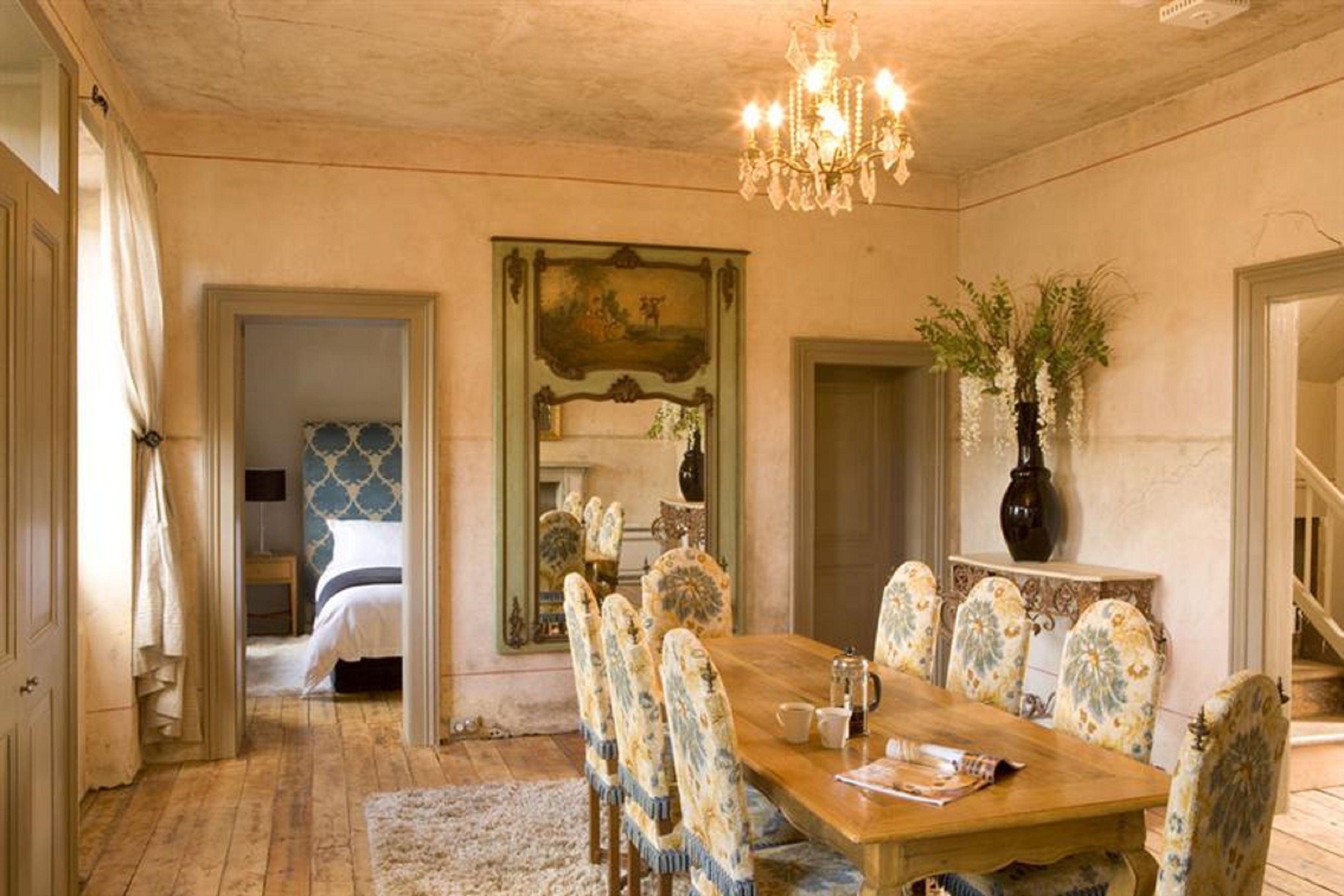 A dining room with a wooden table and floral chairs, a chandelier, and a painting on the wall.