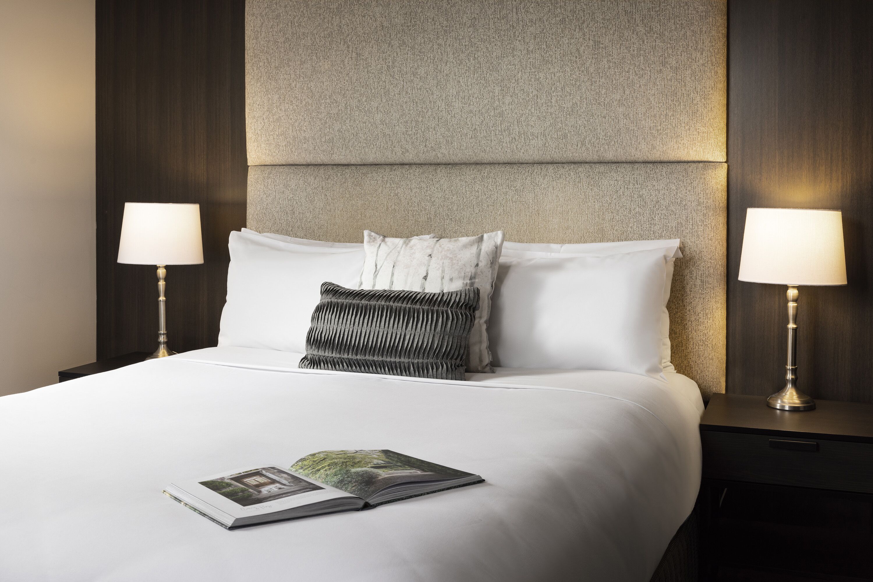 Een net opgemaakt bed met witte lakens, twee bedlampjes en een boek op het bed.