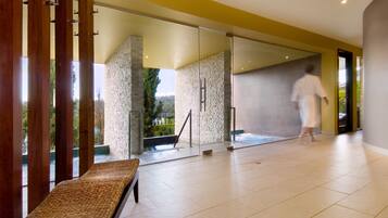 A modern spa area with a glass wall, a wooden bench, and a person in a robe walking towards a door.