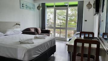Habitación de hotel con cama, sofá, mesa y silla. Balcón con vistas a árboles y edificios.