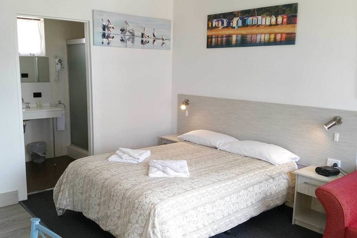 A hotel room with a bed, a nightstand, a red chair, a painting on the wall, and a bathroom visible through an open door.