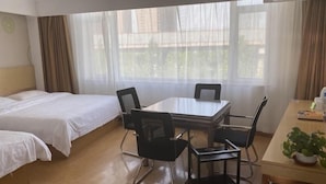 Habitación de hotel con dos camas individuales, una mesa de comedor con sillas, una mesita con una planta y amplias ventanas con cortinas translúcidas.