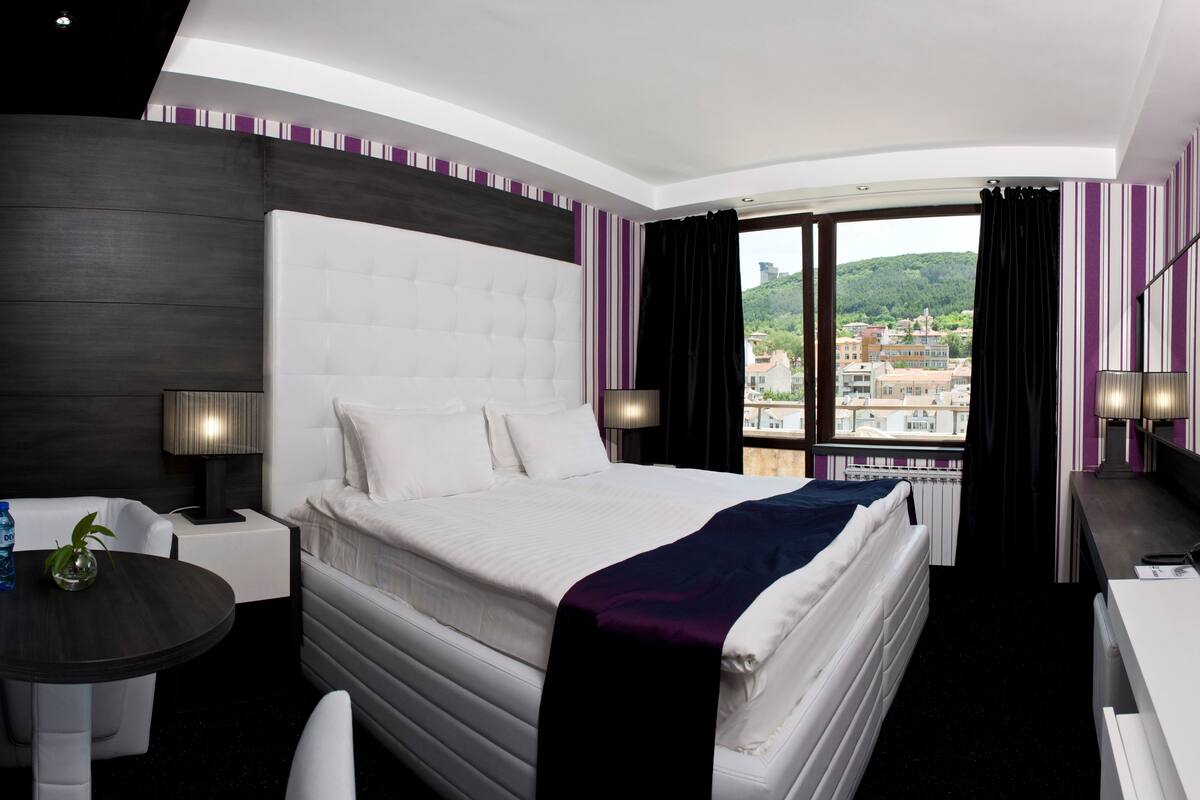 A hotel room with a large bed, a desk, a chair, a TV, and a window with a city view.