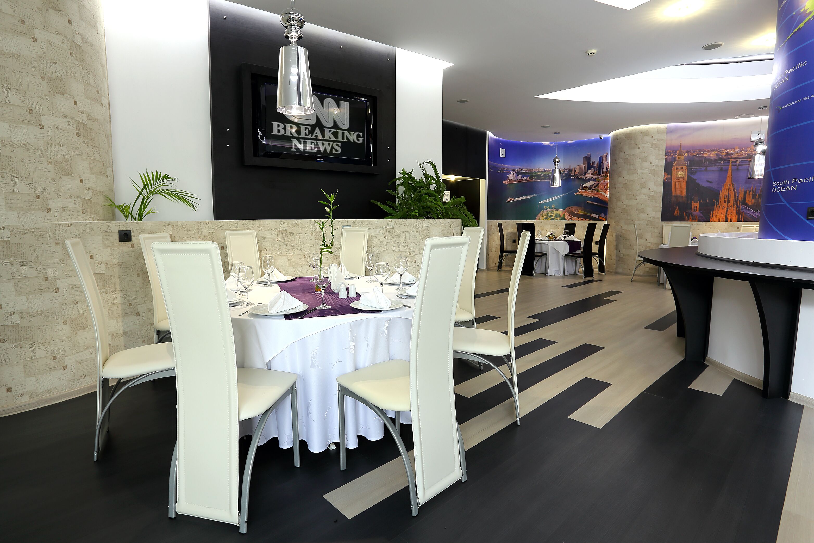 A modern dining area with white chairs and tables, a 'Breaking News' TV screen, and a South Pacific Ocean mural.