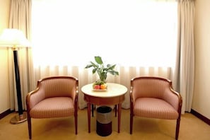 Una habitación de hotel con una mesa redonda, dos sillones, una planta en maceta y una ventana con cortinas.