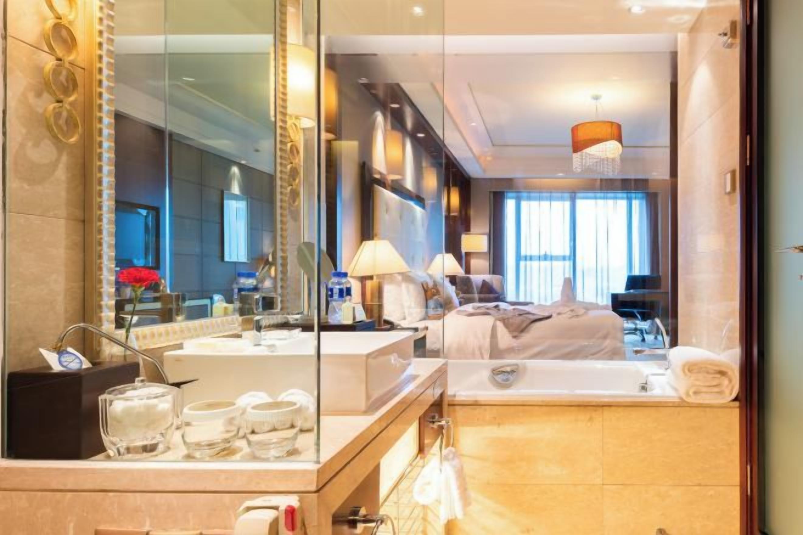 A modern hotel room with a large mirror, a bathtub, a vanity with toiletries, and a bed with a headboard.