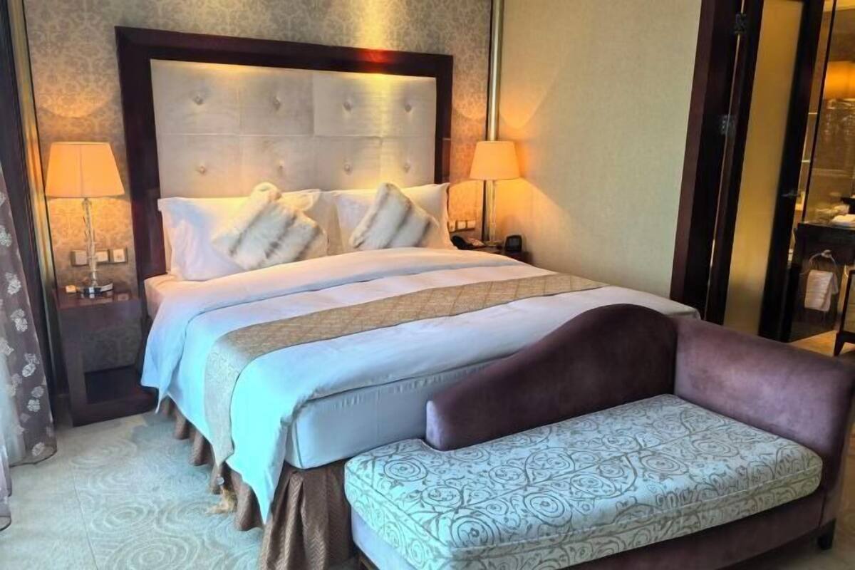 Ein Hotelzimmer mit einem großen Bett, zwei Nachttischen, einer Ablagefläche, einem Sofa und einem gemusterten Hocker.