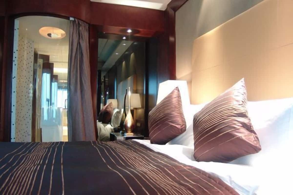 Ein Hotelzimmer mit einem großen Bett, zwei Kissen, einem Nachttisch, einer Lampe und Blick auf den Flur.