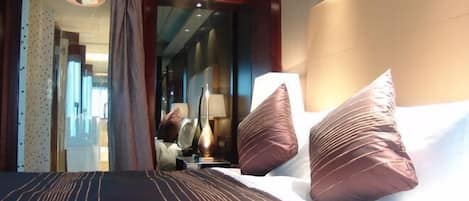 Habitación de hotel con una cama grande, dos almohadas, una mesita de noche, una lámpara y vistas al pasillo.