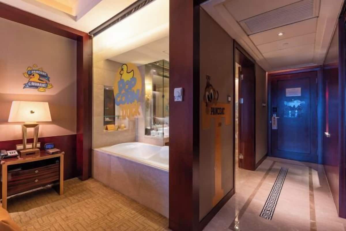 Ein Hotelzimmer mit Bett, Nachttisch, Lampe, Badewanne und einem Flur mit Tür.