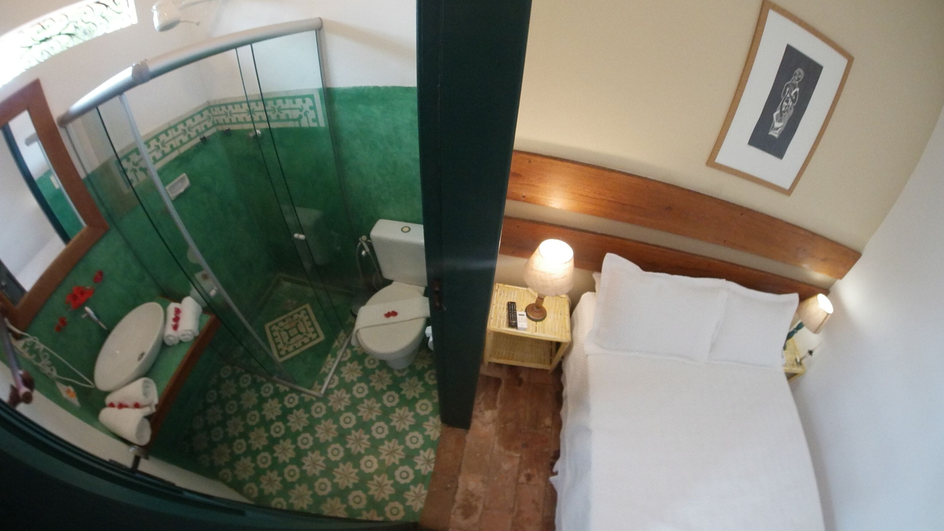 Une salle de bain équipée de toilettes, d’un lavabo et d’une douche. On y trouve un lit avec une tête de lit en bois et une table de chevet avec une lampe.