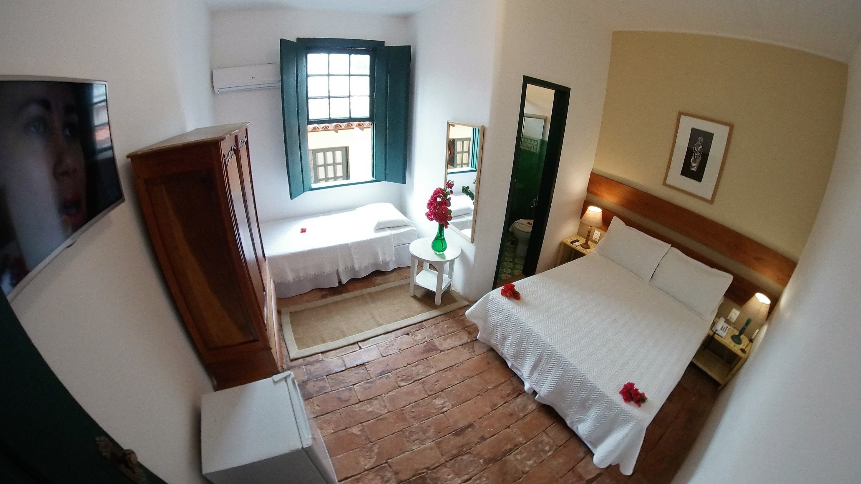 A hotel room with a bed, a small table with a vase of flowers, a window with curtains, a mirror, and a television.