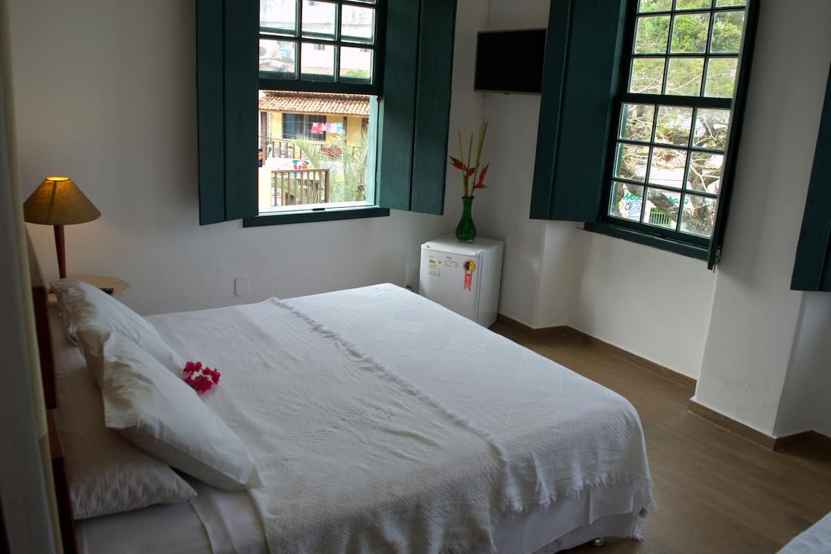 Un dormitorio con cama, mesita de noche, lámpara, un florero verde con flores y dos ventanas con contraventanas verdes.