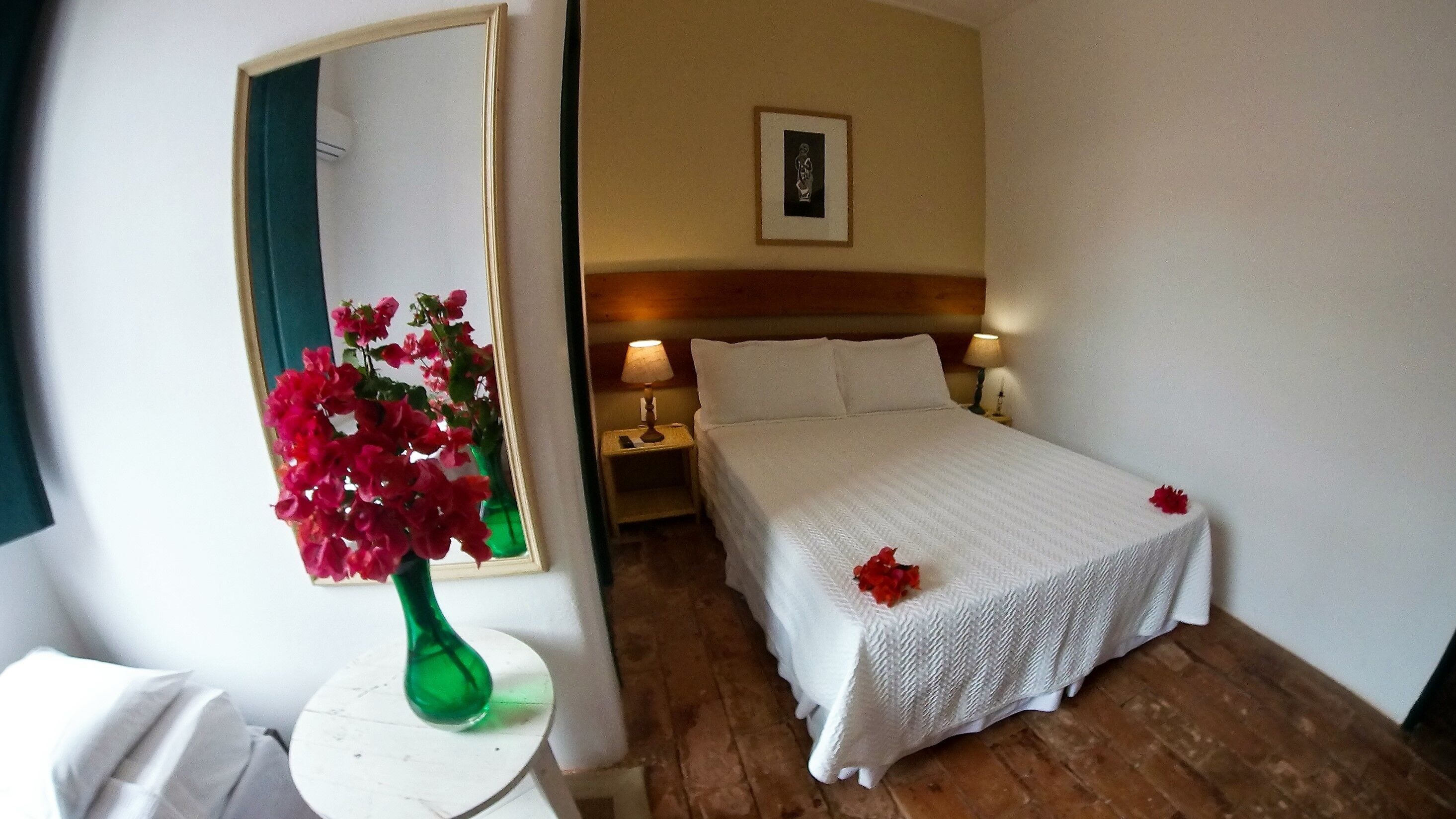 A hotel room with a bed, a nightstand, a lamp, a mirror, and a vase with flowers.