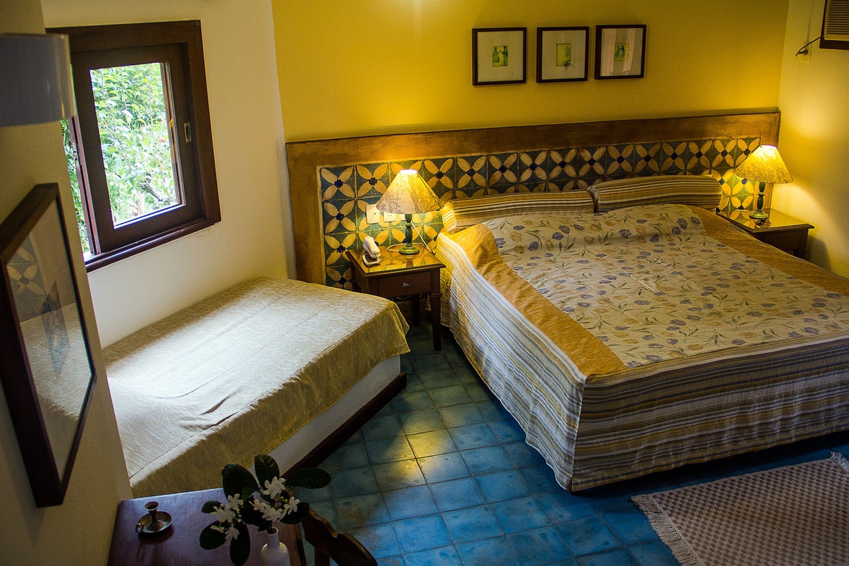 A room with a bed, a bedside table with a lamp, a small table with a vase, and a window.