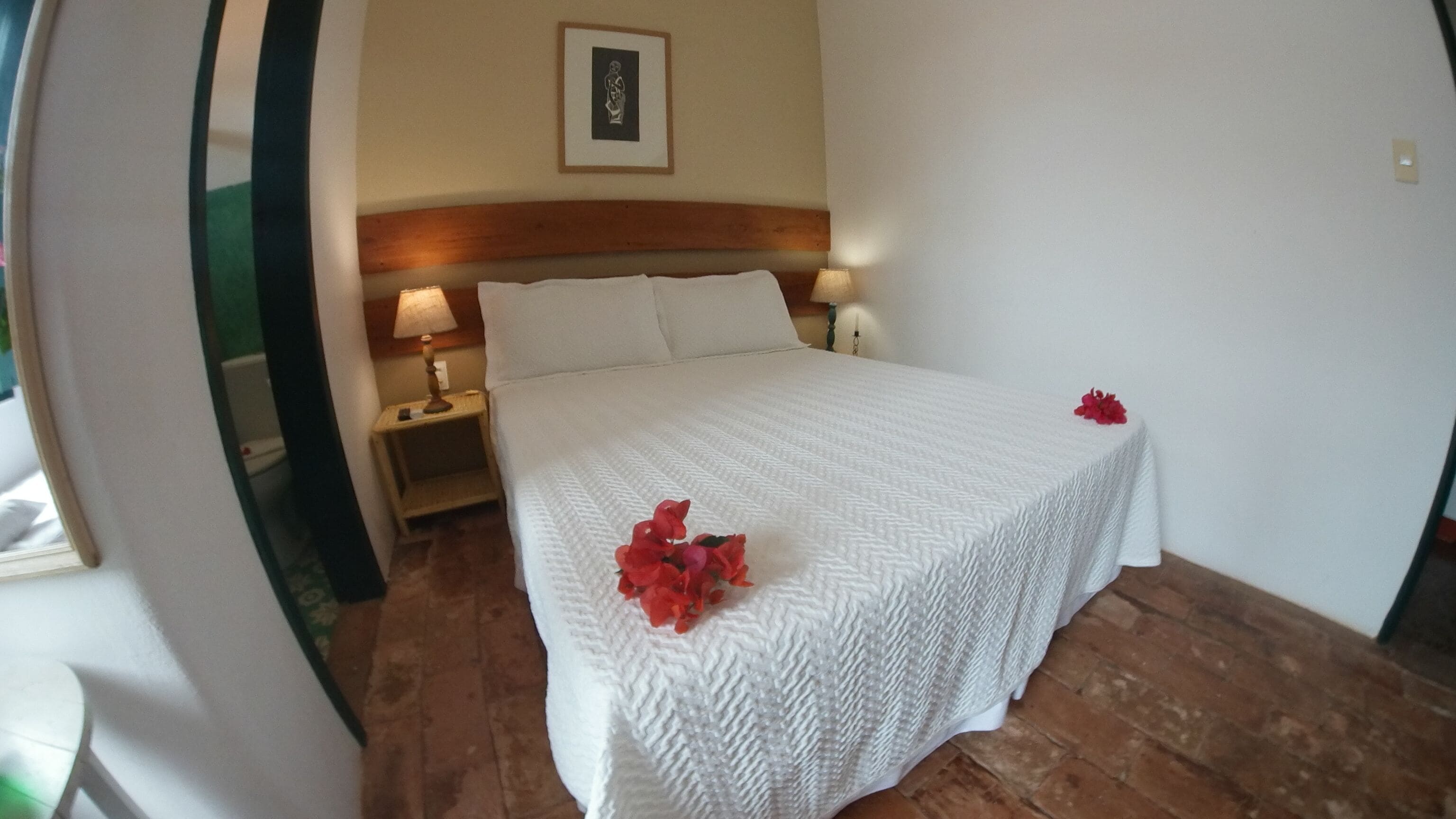A neatly made bed with a white bedspread and two red flowers on the side. There is a wooden headboard, a nightstand with a lamp, and a framed picture on the wall.