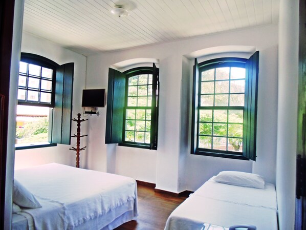 A room with two beds, large windows with green shutters, a flat-screen TV mounted on the wall, and a wooden coat rack.