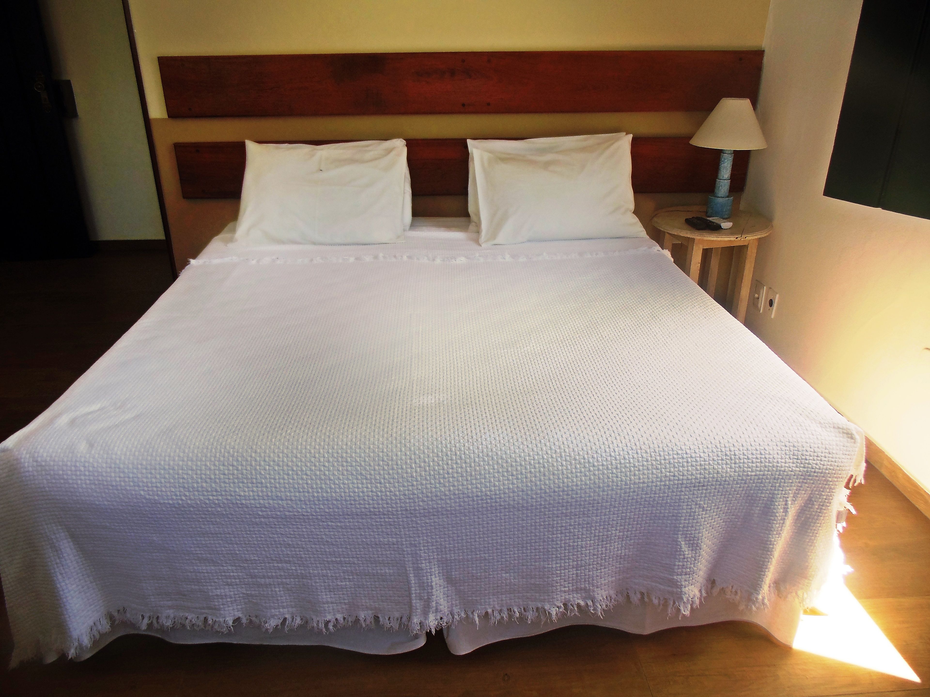 Een net opgemaakt bed met witte lakens, een houten hoofdbord en een klein nachtkastje met een lamp.
