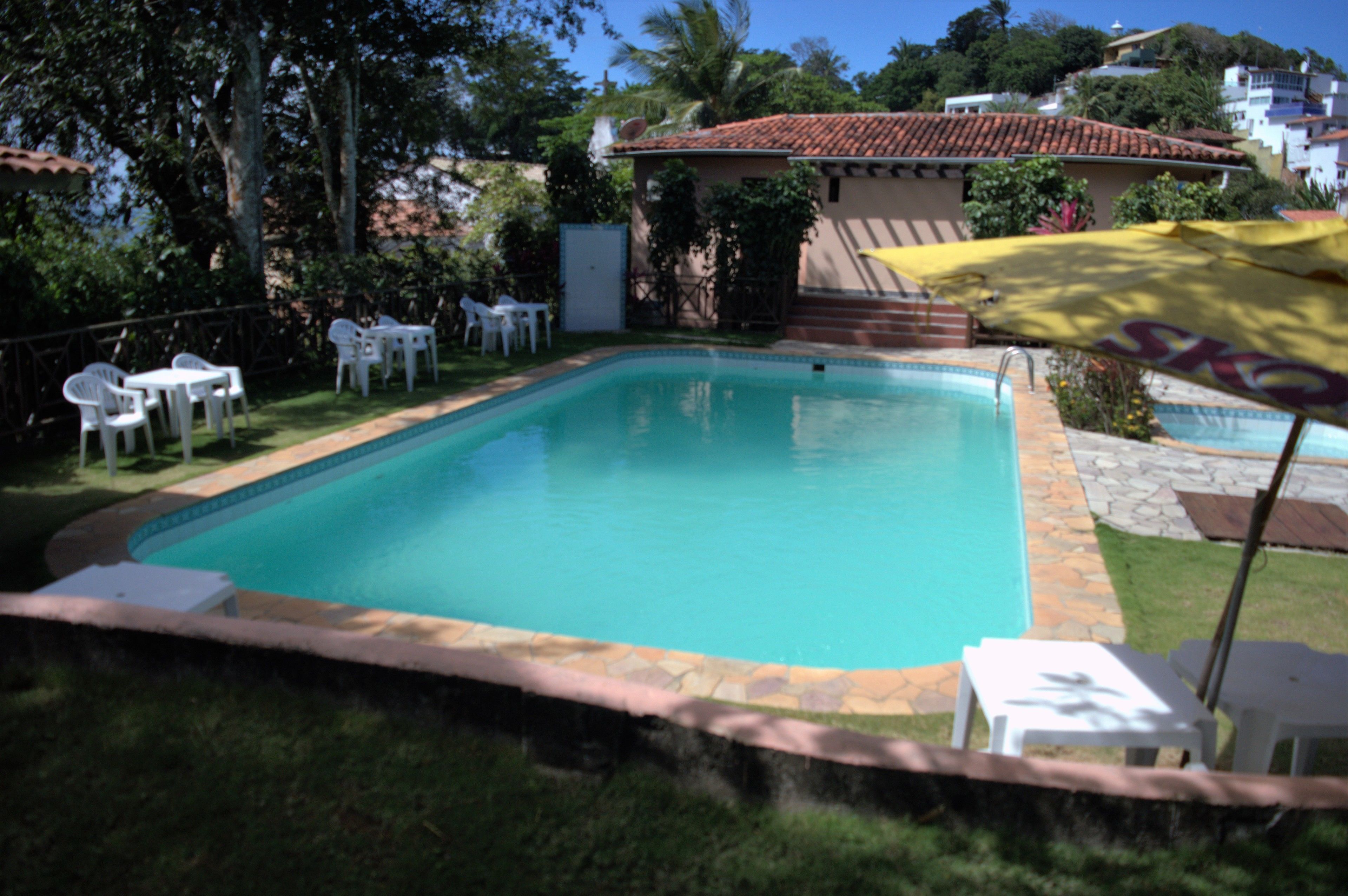 Una piscina rectangular con borde de azulejos, rodeada por un marco de piedra. Se observan sillones blancos y un paraguas amarillo a la derecha.