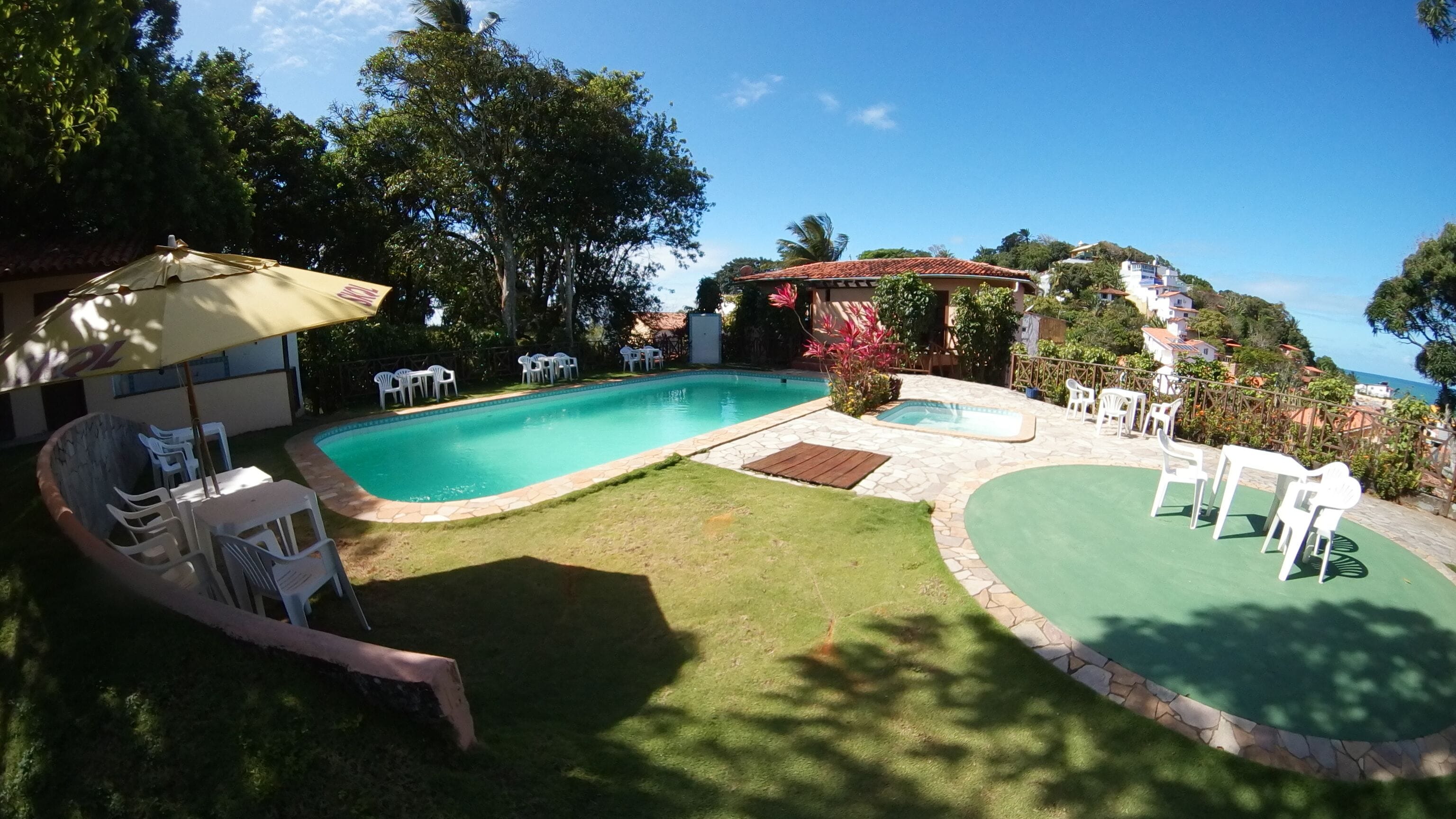 A swimming pool with a stone border, surrounded by grass and trees. There are white plastic chairs and a table under an umbrella.