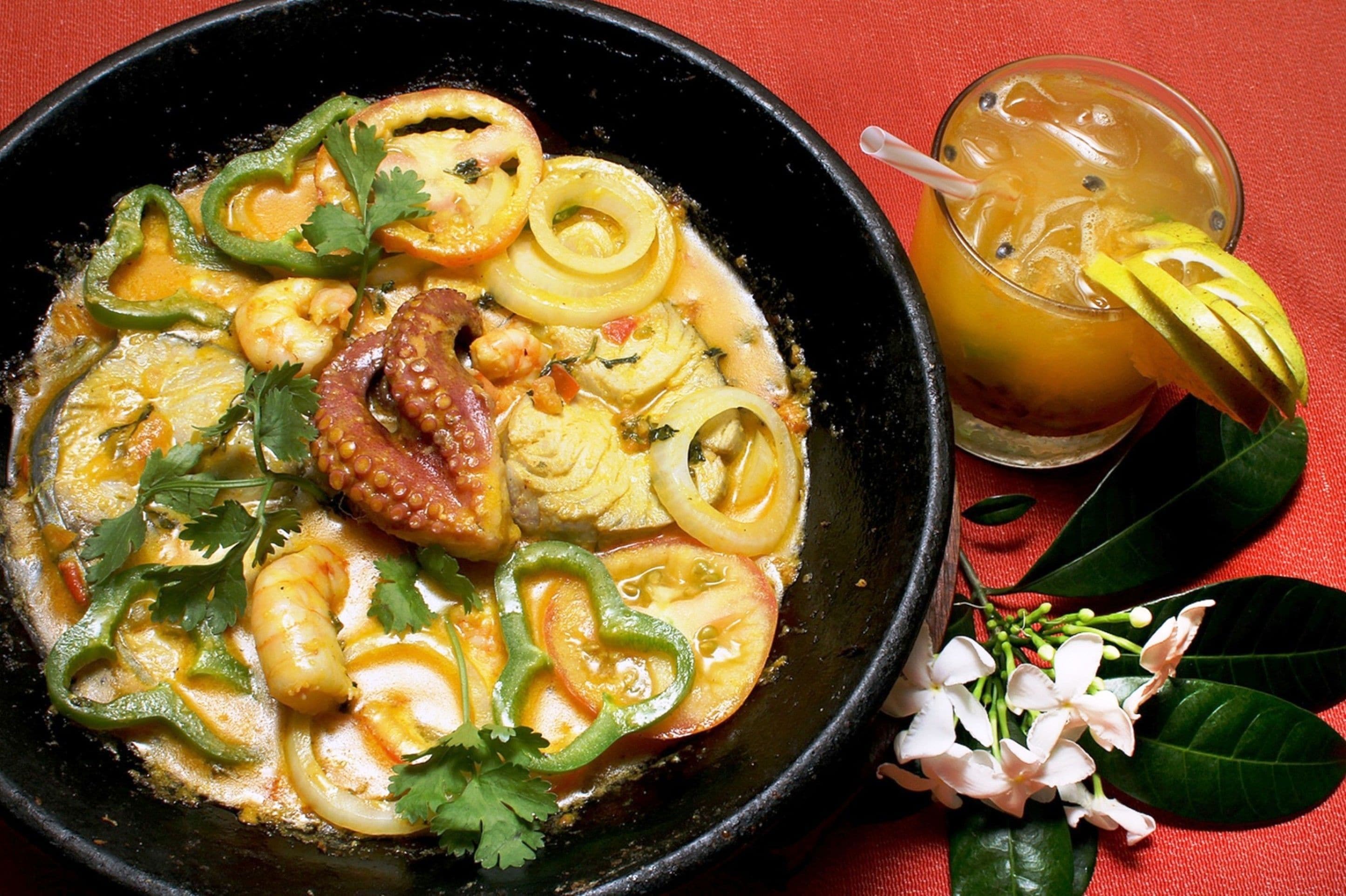 A bowl of seafood curry with sliced vegetables and a glass of iced drink with a lemon slice on the side.