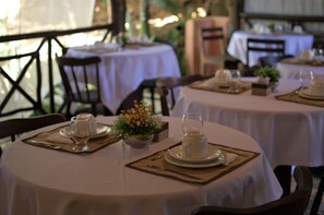 Un comedor con mesas redondas dispuestas para dos comensales, cada una con mantel blanco, un pequeño centro de mesa floral y cubiertos y platos ordenados.