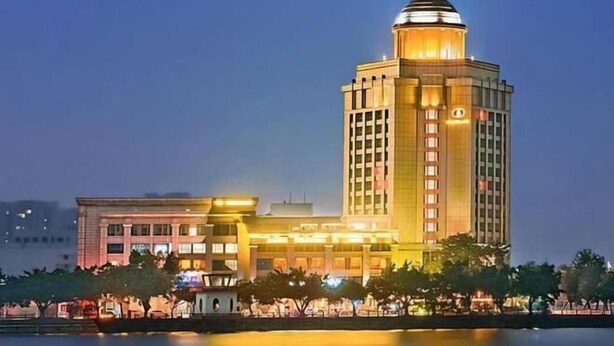 A large, multi-story building with a prominent dome on top, likely a hotel or corporate building, with a well-lit facade and a reflection in the water in front. - Sanbu Holiday Hotel (Jiangmen)