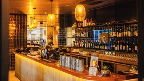 A bar area with a wooden counter, hanging light fixtures, and shelves stocked with bottles. - Shoreline Hotel (Howrah)